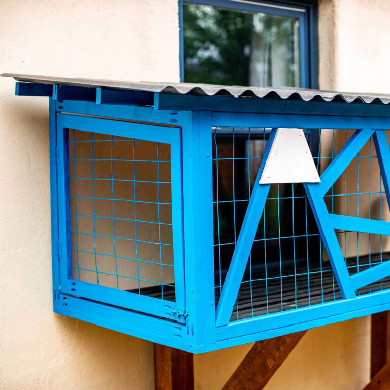 Close-up side view of a small bright blue catio with wire mesh, a pitched corrugated roof, and triangular decorative elements, mounted outside a house window.