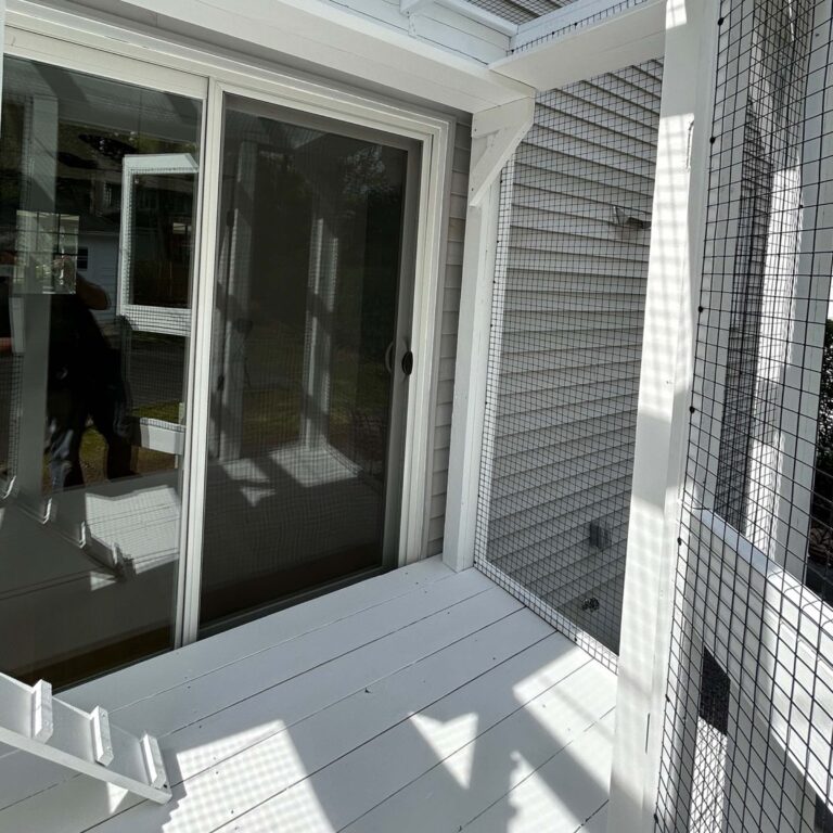 Custom white-painted small catio structure attached to a house with a sliding glass door, wire mesh walls, and partial sunlight casting shadows on the wooden floor.