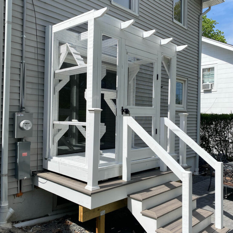 Angled side view of a small white catio with a peaked roof structure, mesh walls, wood stairs, and multiple platforms inside for cat climbing.