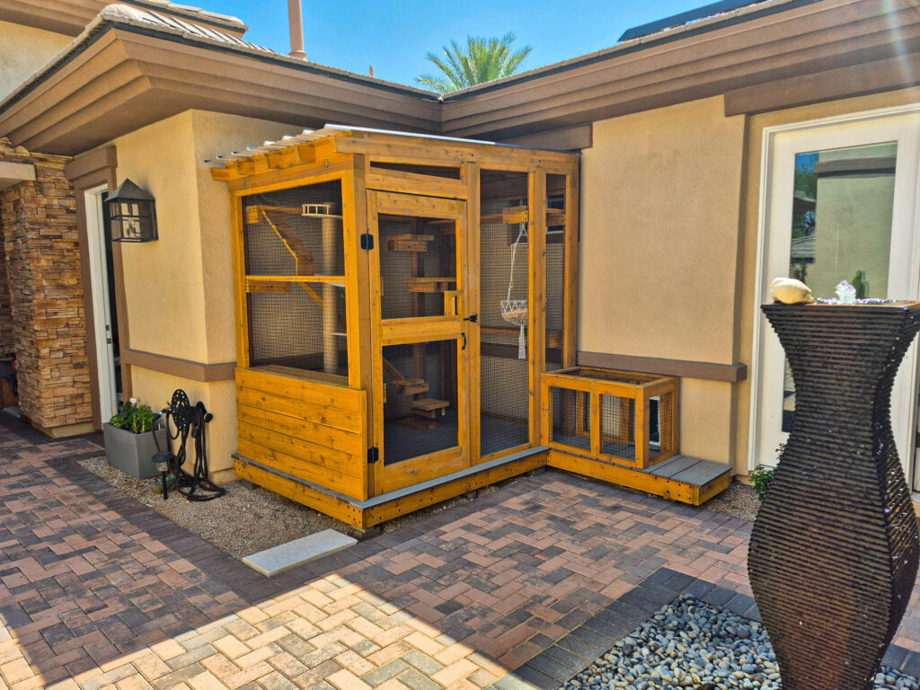 Corner-mounted wooden catio with climbing shelves and a window entry, seamlessly integrated into a backyard patio with stone and tile accents.