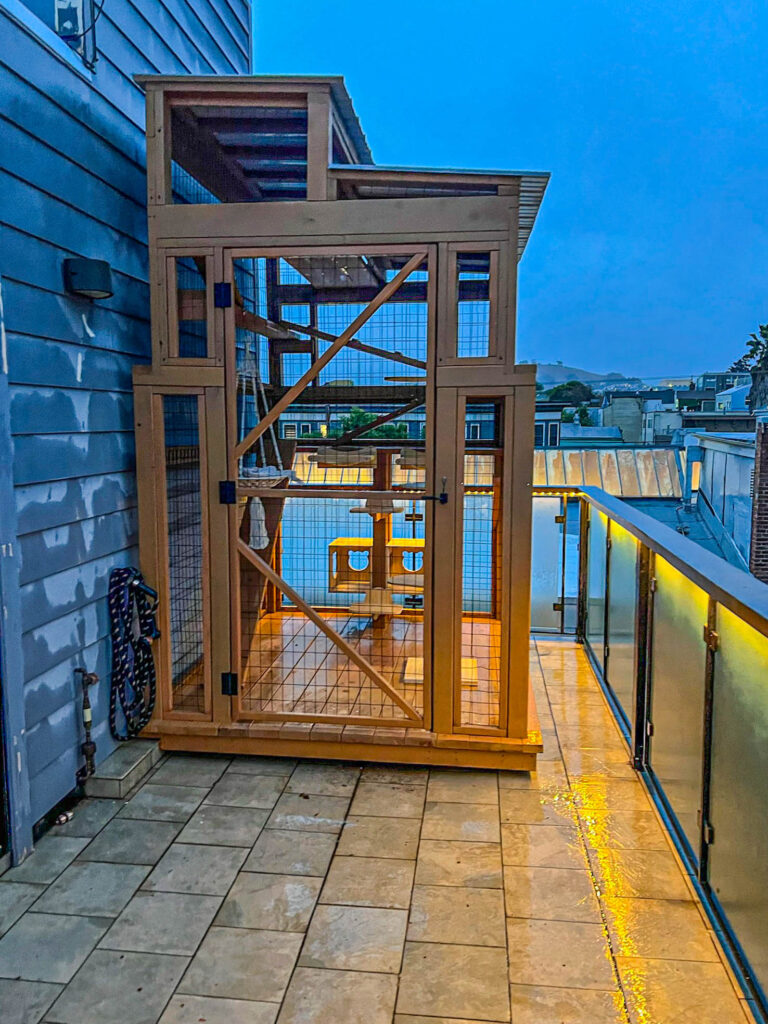 Tall custom catio built on a balcony with vertical climbing ramps, enclosed with wood framing and wire mesh, overlooking an urban sunset.
