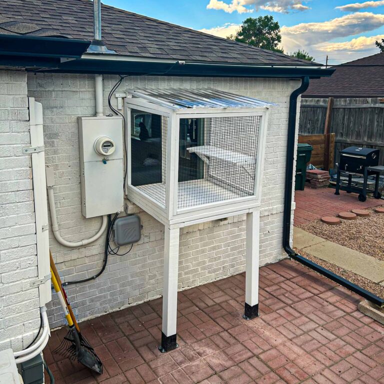 Angled view of a tall, white framed window catio with mesh sides and a clear roof, securely mounted to a white brick wall above a brick patio.
