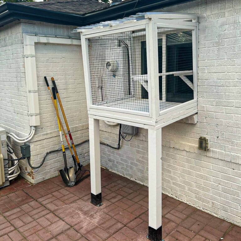 Outdoor side view of a small white window-mounted catio with a clear polycarbonate roof and tall support legs on a brick patio.