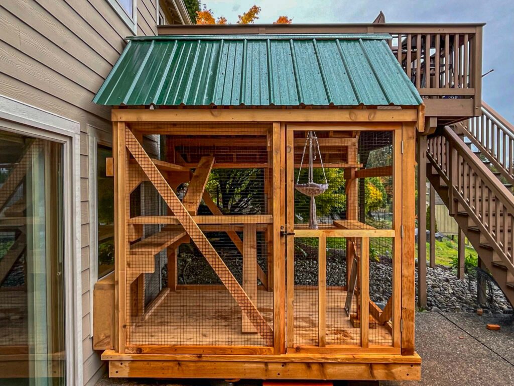 Multi-level outdoor catio with a green metal roof, wooden climbing shelves, and a macrame cat hammock, attached to the side of a house.