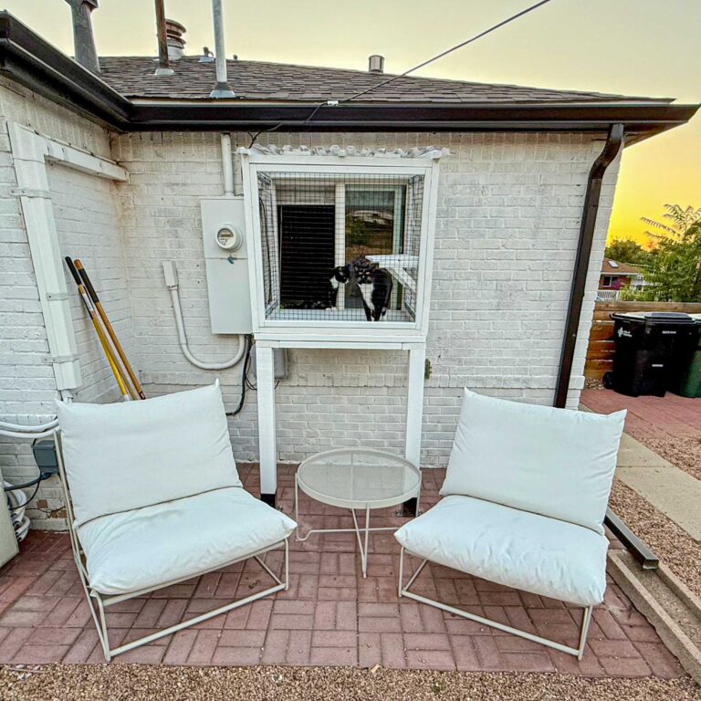Black and white cat peering through a small elevated window box catio attached to a white brick house, with two white patio chairs and a round glass table in front.