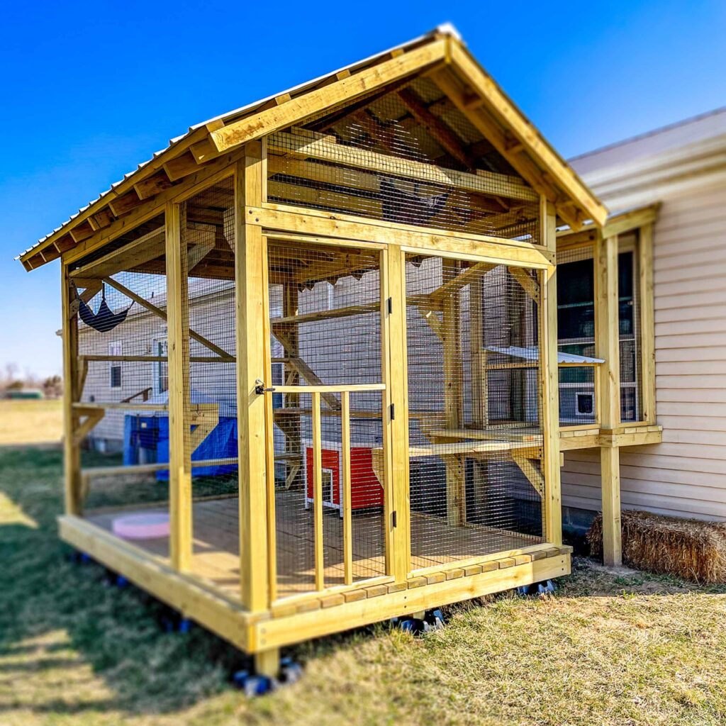 Large wooden custom catio with a gabled roof connected to a house, featuring hammocks and climbing shelves.