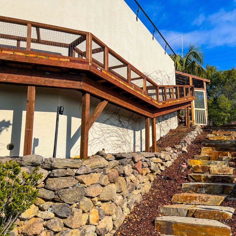 Elevated catio tunnel running along a stone retaining wall beside a modern home.