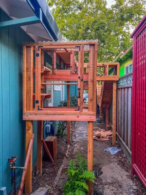 Elevated custom catio with front-facing mesh door and secure wooden frame attached to a teal house exterior