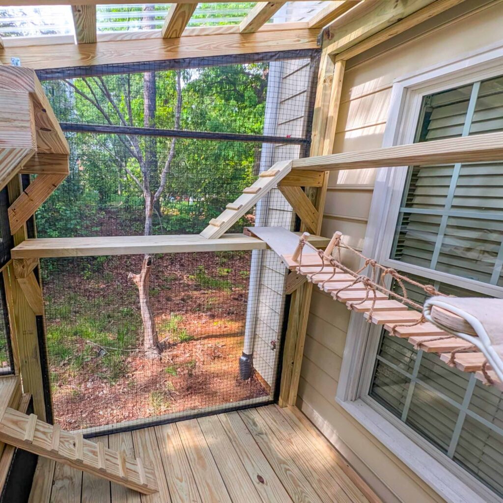 Corner view of a wooden catio with angled climbing ramps, flat bridge, and rope walkway connecting two walls.