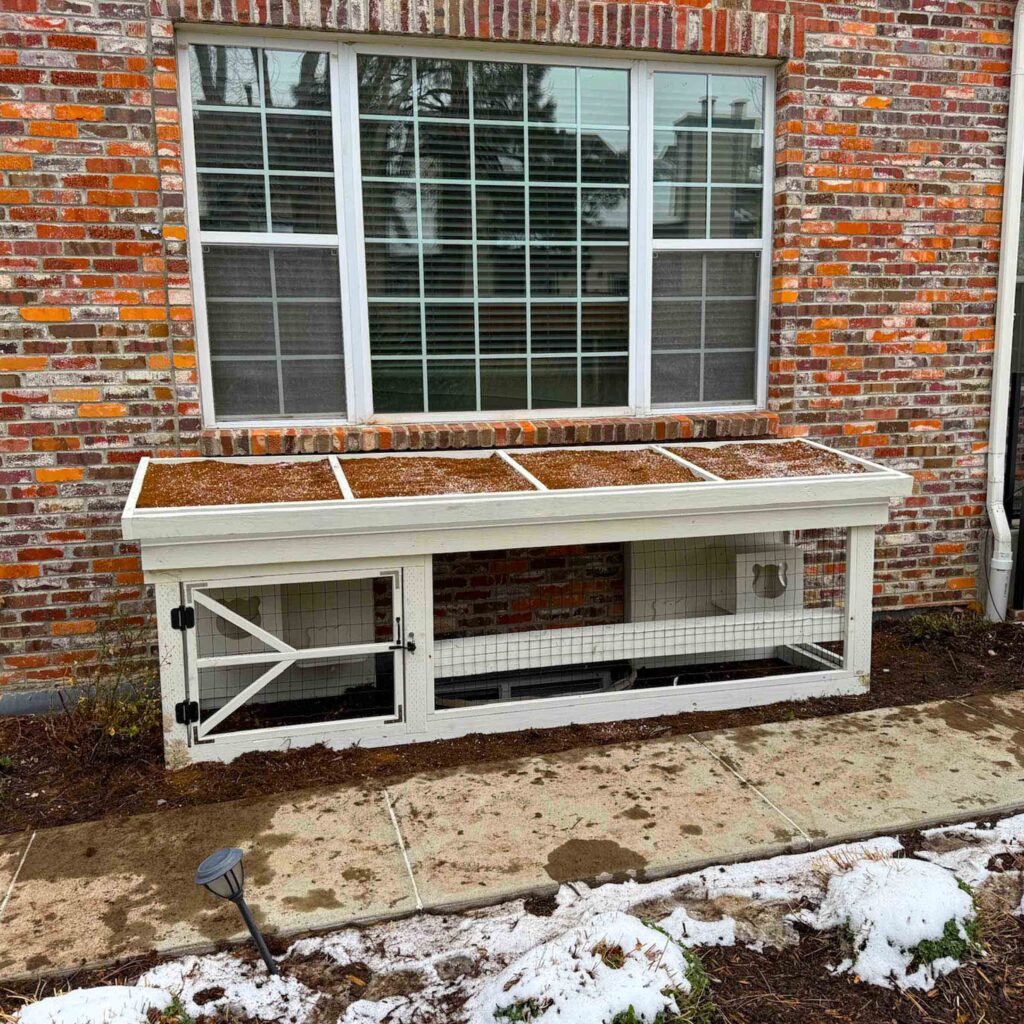 Low-profile white catio attached to a brick house with a window access point.