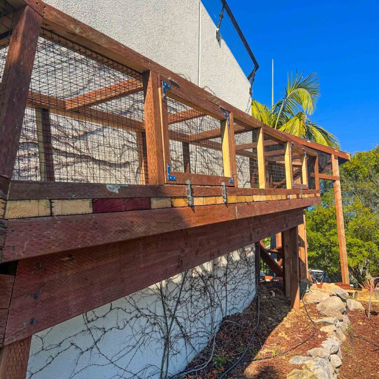 Long wooden elevated catio tunnel attached to the side of a house, supported by posts and framed with mesh sides.