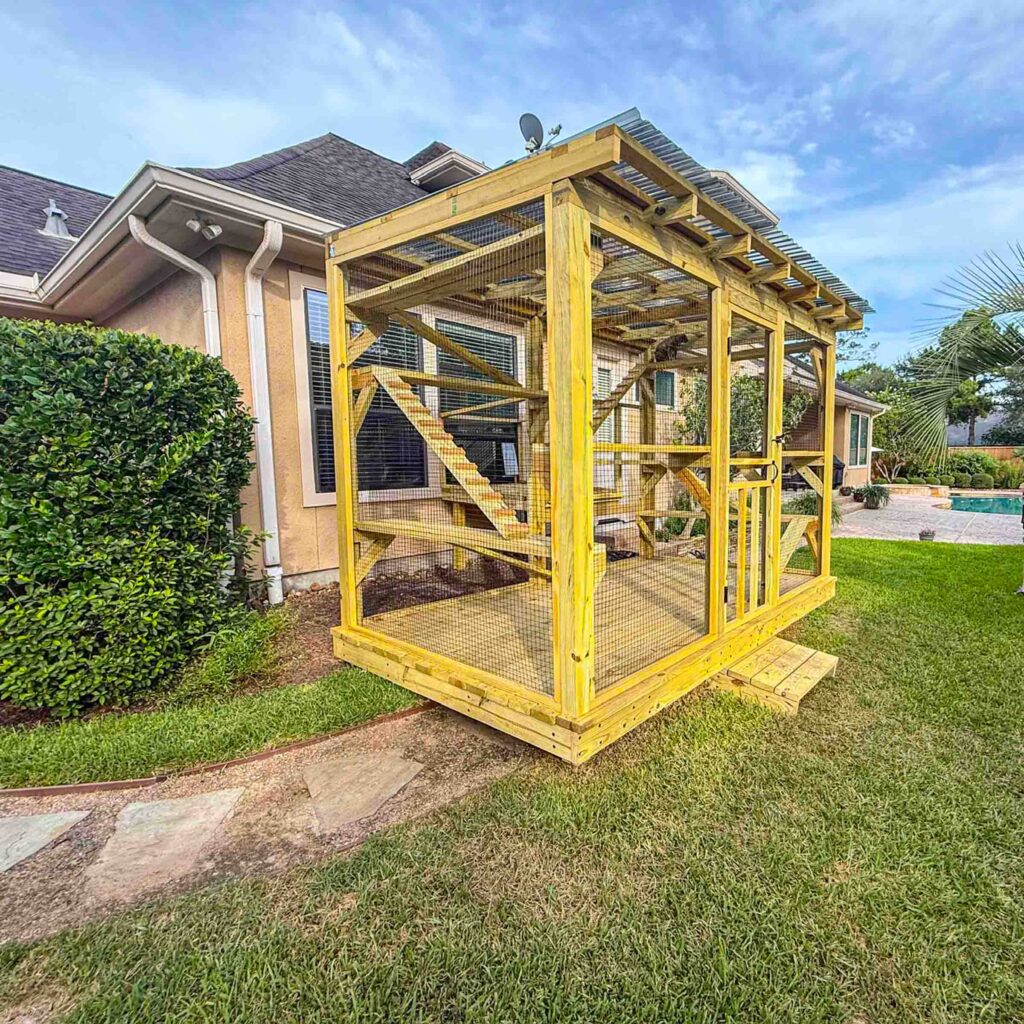 Outdoor catio built with light wood frame, mesh enclosure, and elevated platforms beside landscaped garden