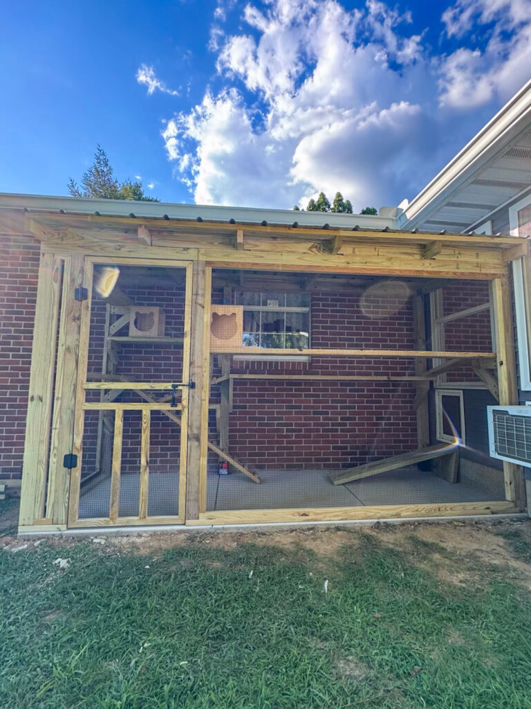 Custom outdoor catio attached to a red brick home in Sevierville, Tennessee, featuring a wooden frame, screened walls, multiple climbing platforms, and a secure entry door.