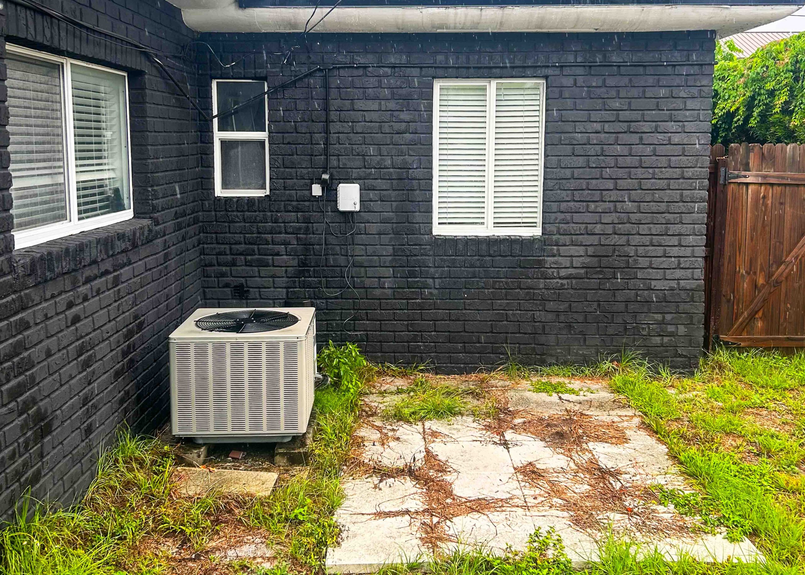 Backyard space before catio installation showing a small patio area with concrete pavers, an air conditioning unit, and dark brick exterior walls.