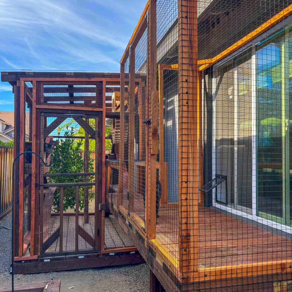 Long custom-built wooden catio running alongside a home with multiple sections, secure doors, and mesh panels providing outdoor access through sliding glass doors.