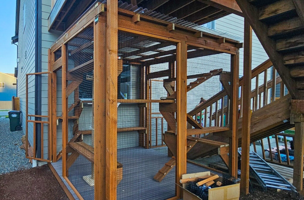 Side view of a custom-built catio in Yelm, WA, showing sturdy cedar framing, wire mesh walls, and elevated platforms for climbing beneath a wooden staircase.