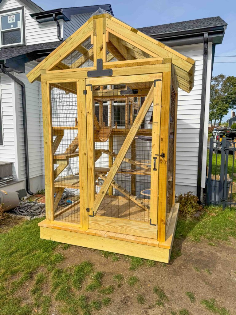 Custom wooden catio with a pitched roof and secure wire mesh, located outside a white house in Weymouth, Massachusetts.