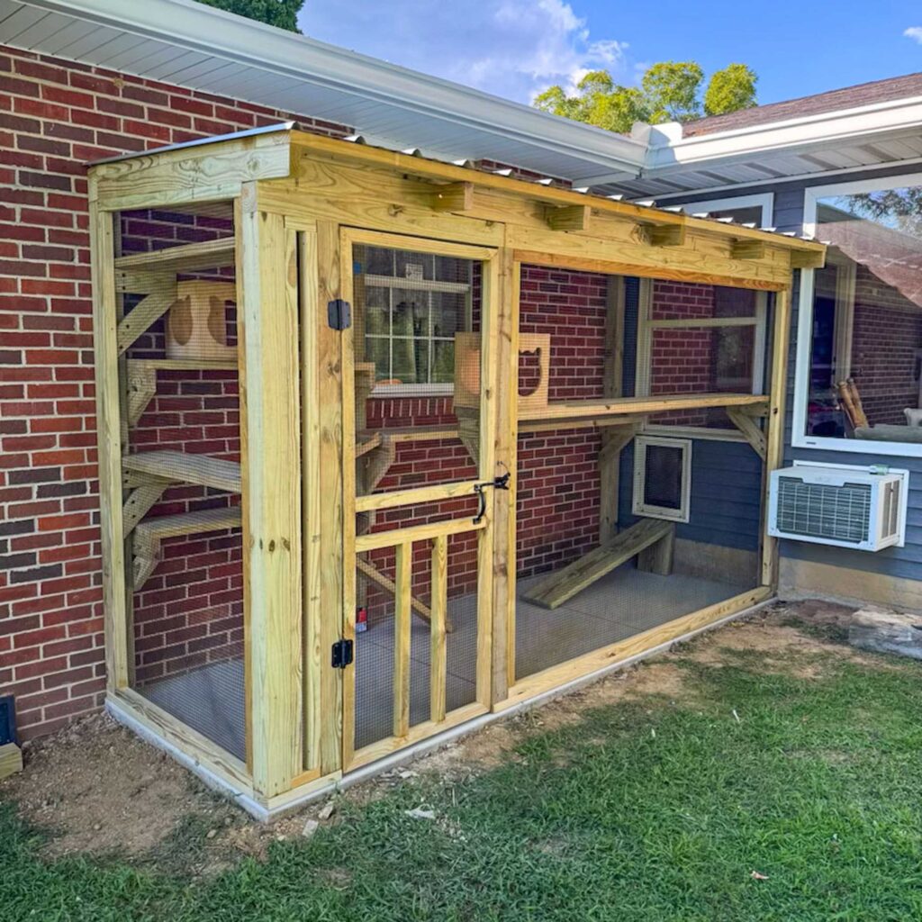 Long wooden catio attached to a brick home with shelves and climbing structures inside.