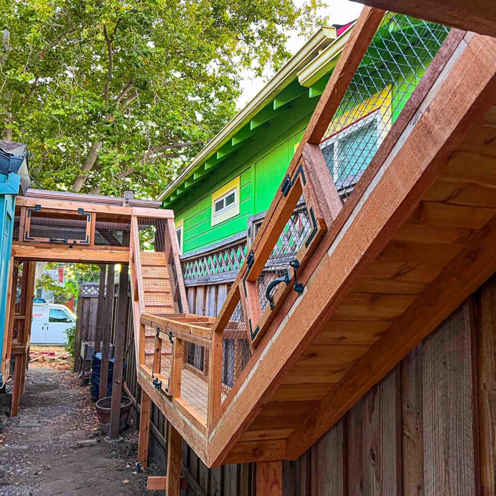 Multi-level catio built along a colorful house exterior with wood framing, mesh panels, and angled enclosures leading up to a bridge connecting two sections.