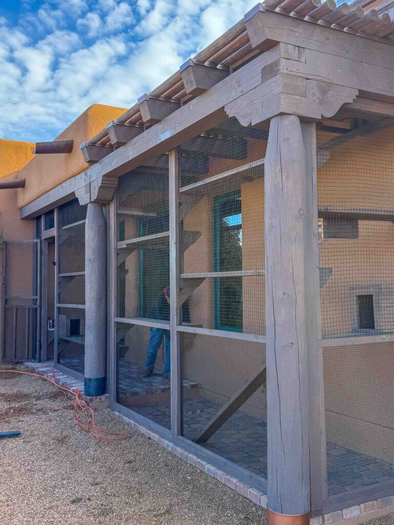 Exterior view of a custom-built catio in Santa Fe, New Mexico with wood framing, secure mesh panels, and adobe-style architecture under a partly cloudy sky.