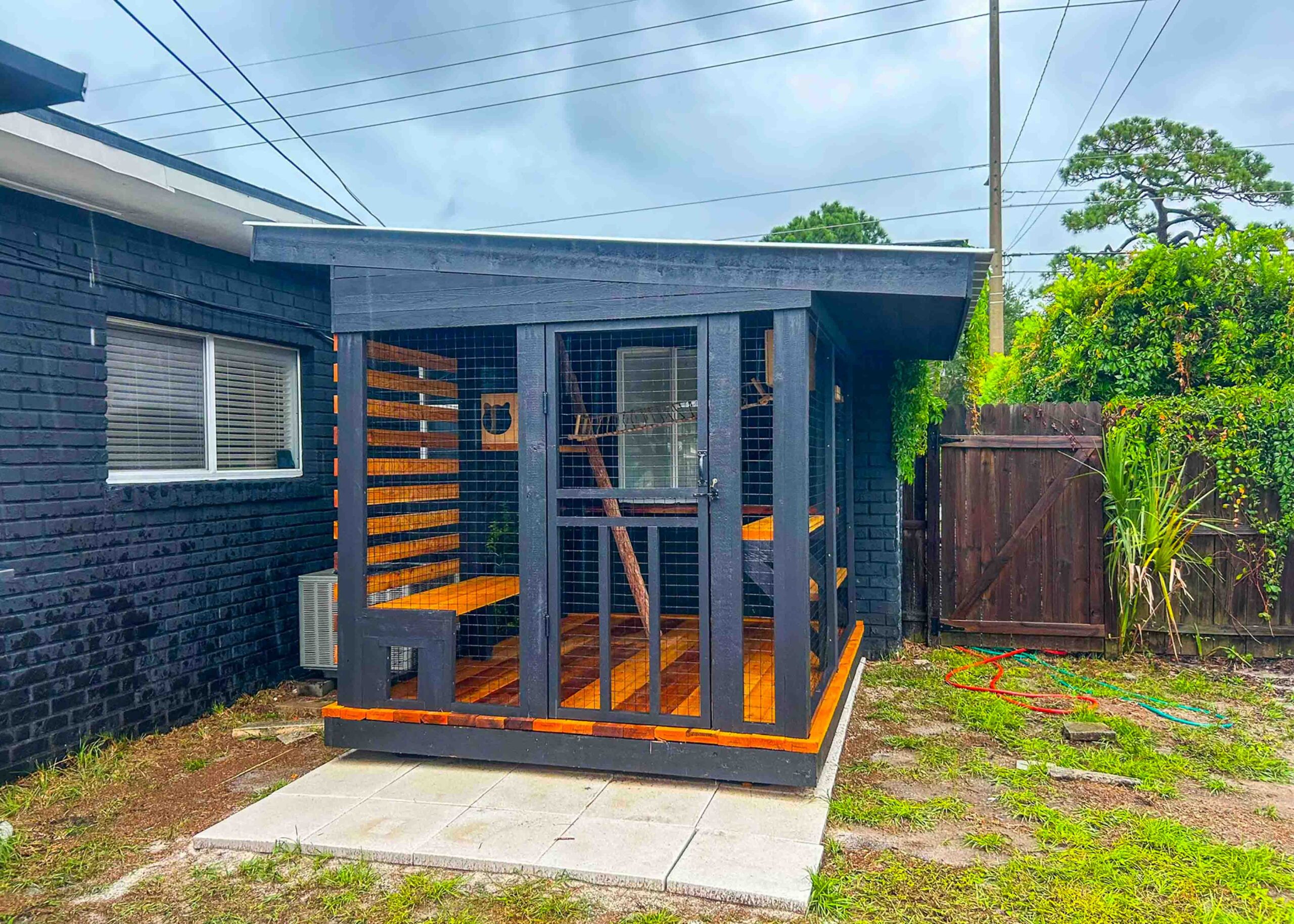 Modern black catio enclosure with orange trim built against a dark brick home, featuring a slanted roof and enclosed seating platforms.