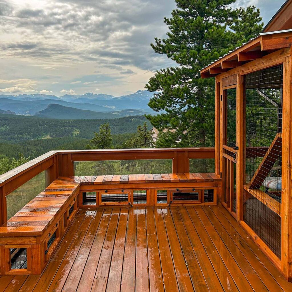 A large wooden catio on a mountain-view deck with matching stained wood benches, surrounded by a breathtaking landscape of rolling hills and distant peaks.