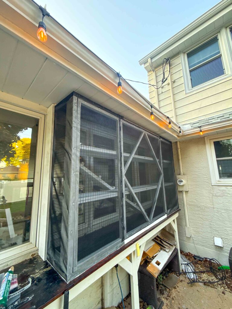 Angled side view of a window catio attached to a beige home in Roseville, MN with black mesh, metal framing, and warm outdoor lighting.