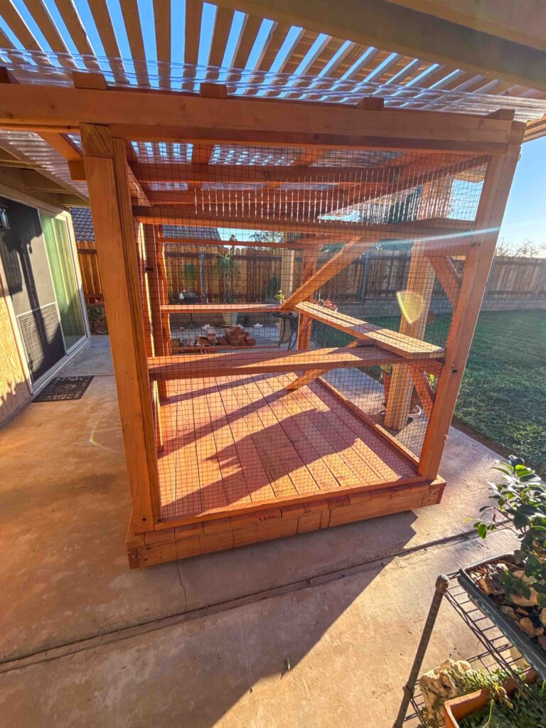 Custom wooden catio in Moreno Valley, California, featuring wire mesh walls, multiple climbing platforms, and a covered roof extension for shade and weather protection.