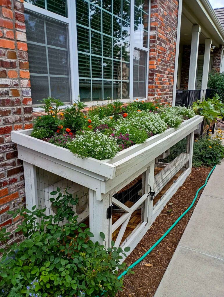Low-profile white window-entry catio installed beneath a large window with a planted flower box on top.