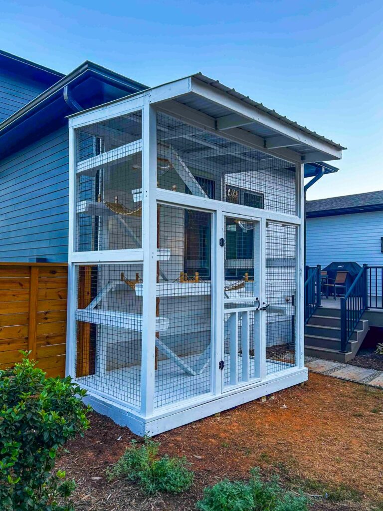 Tall white custom catio with a covered roof, multiple interior platforms, ramps, and hammocks attached to a blue home.