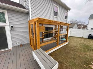 Exterior view of a custom wooden catio attached to a house, featuring a clear roof, wire mesh panels, and built-in climbing platforms.