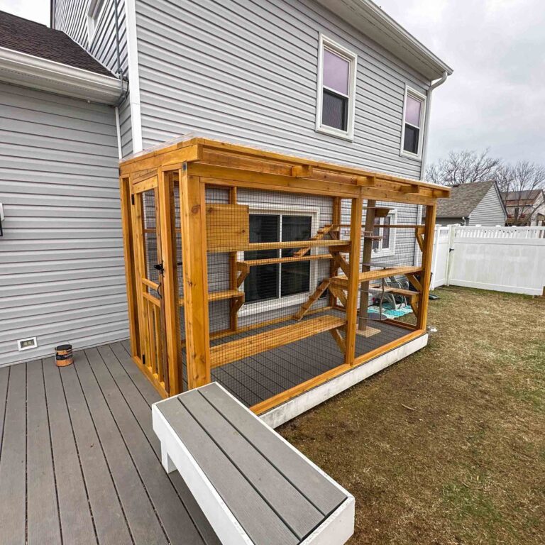 Exterior view of a custom wooden catio attached to a home, featuring wire mesh panels, a sloped roof, and multiple interior climbing levels.