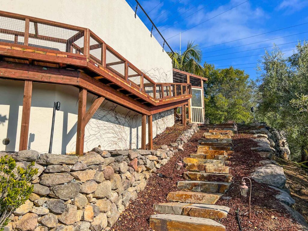 Elevated wooden catio walkway built along a hillside with stone steps and landscaping.