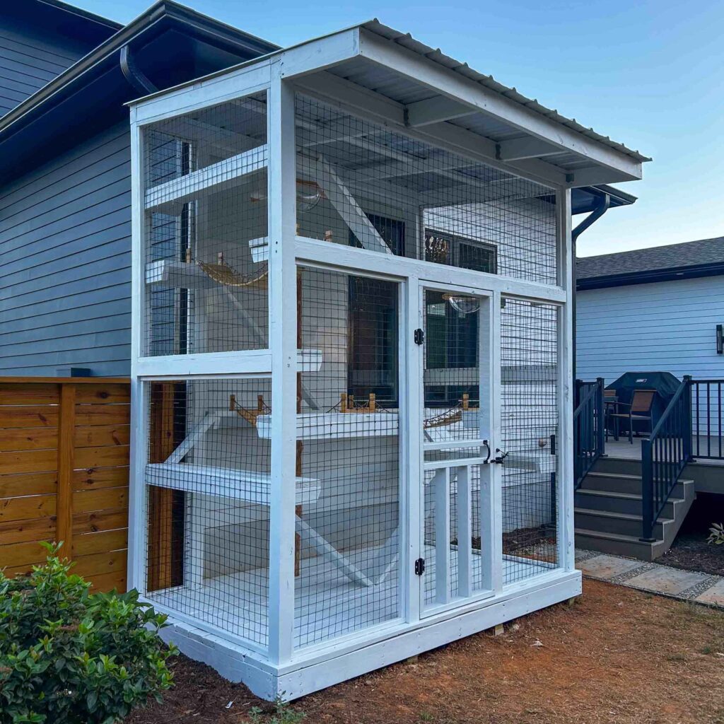 A white custom-built catio with multiple climbing shelves and a wire mesh exterior, shown from a front-left angle.