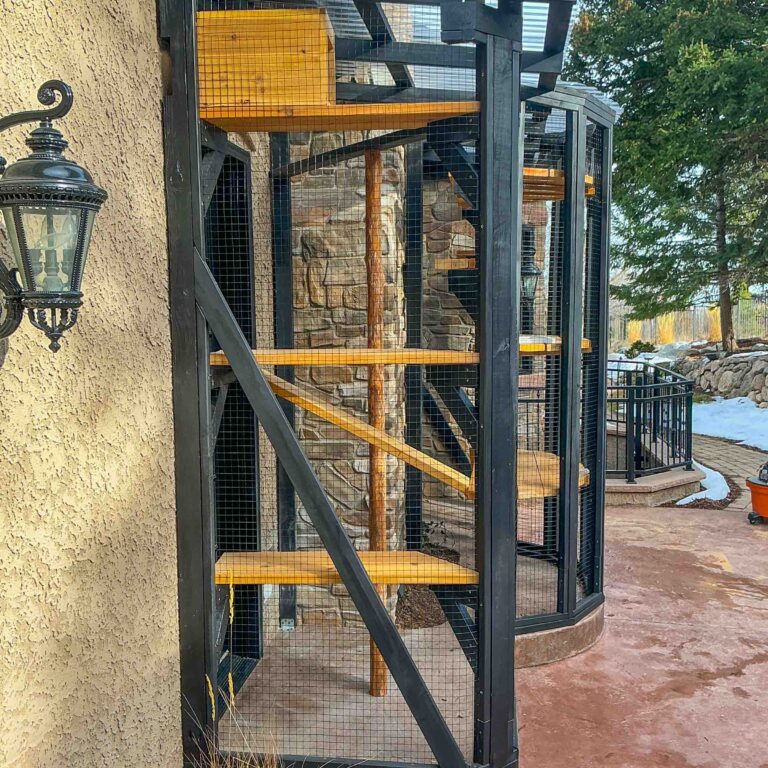 Side view of a tall custom catio with vertical cedar platforms, black structural supports, and wire mesh enclosure along a home exterior.