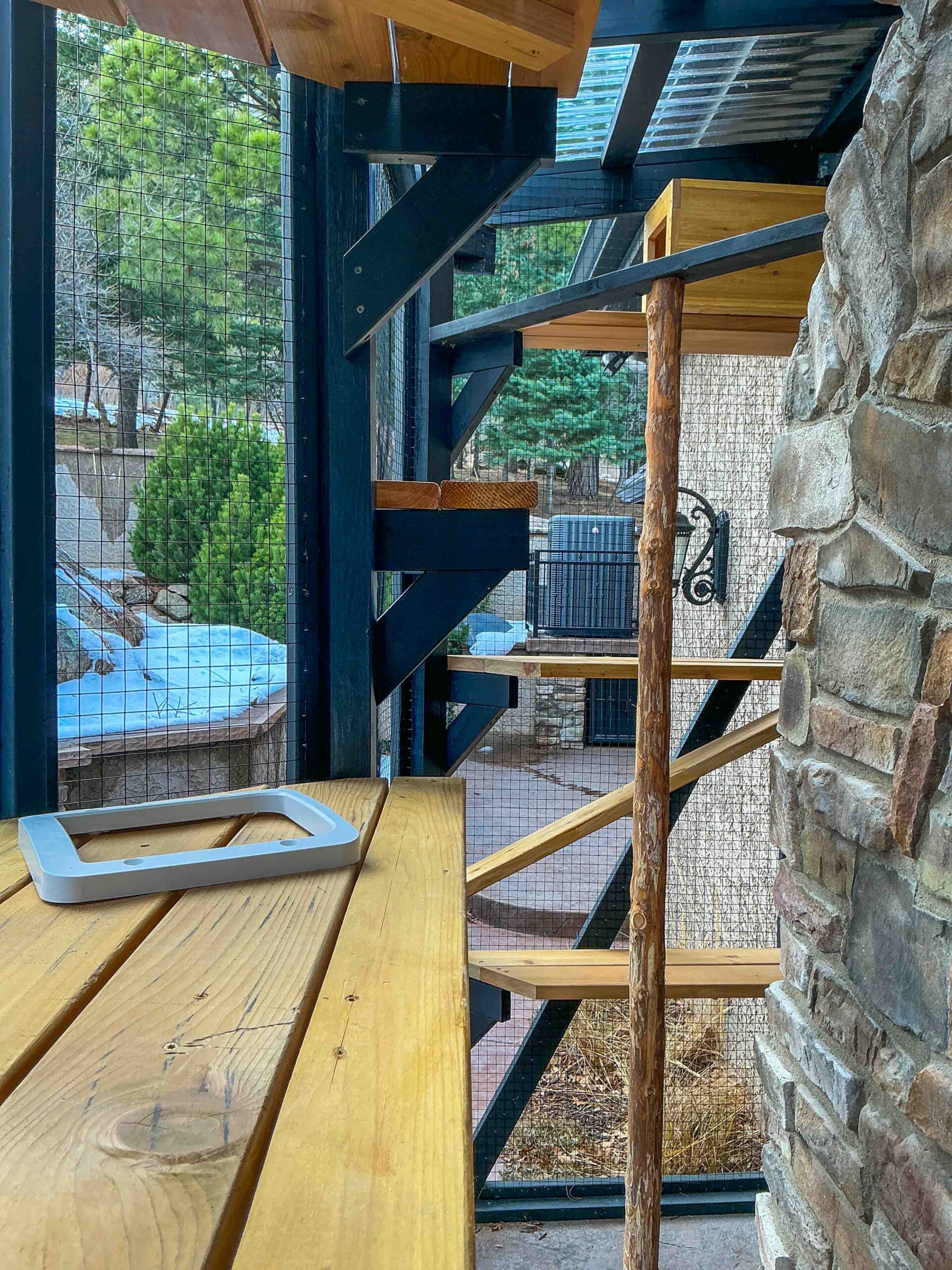 Interior view of a custom catio showing wooden platforms, natural branch supports, and wire mesh walls with an outdoor view.