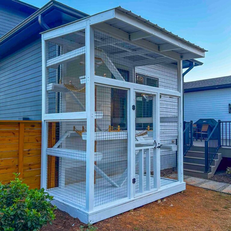 Tall freestanding white catio enclosure with a metal roof, secure wire mesh walls, interior ramps, shelves, hammocks, and a full-height access door installed beside a home.