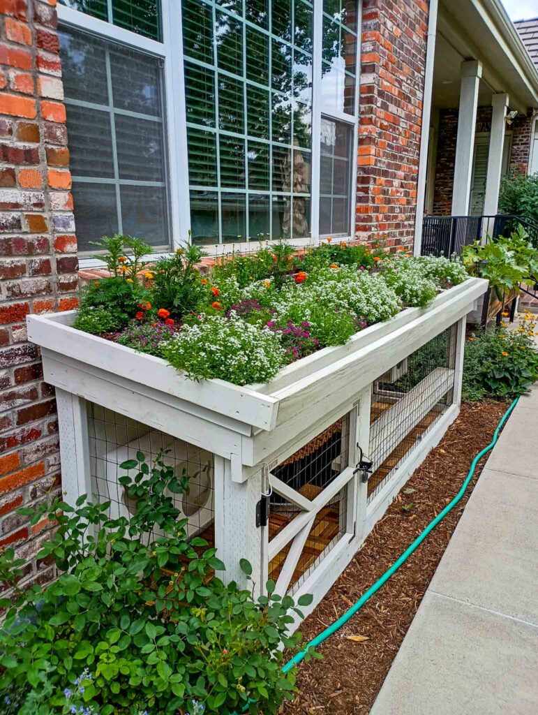 White window catio with a planter box roof filled with flowers built against a brick home.