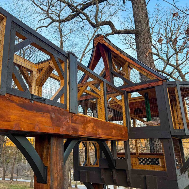 Elevated custom wooden catio tower and connecting catwalk built with rich cedar planks and dark charcoal framing, enclosed in galvanized wire mesh and supported by heavy timber posts among mature backyard trees.