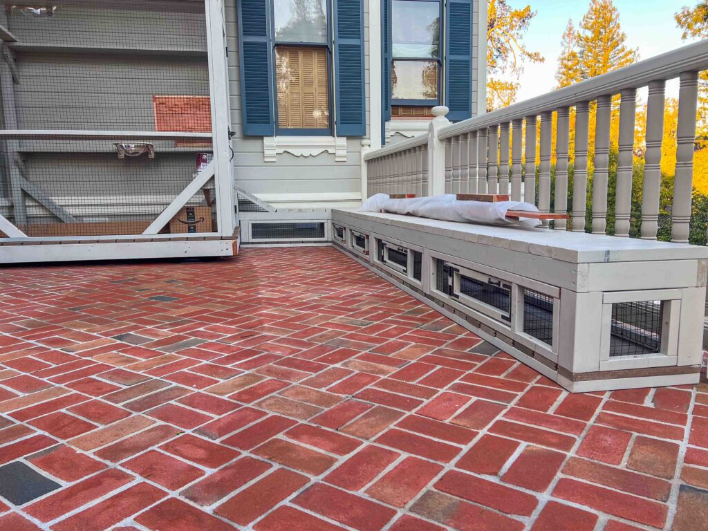Low-profile custom catio bench enclosure built along a porch railing in Atherton, California, featuring integrated wire-mesh access panels beneath a long cushioned lounging surface on a red brick patio.