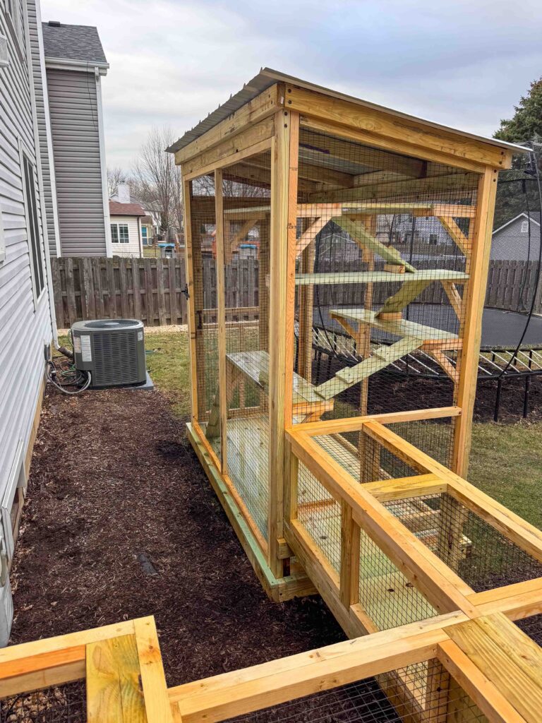 Custom-built wooden catio in Carol Stream, IL attached to the side of a home, featuring a fully enclosed wire mesh structure with a slanted corrugated metal roof, elevated foundation, and an attached tunnel extension leading from the house to the main enclosure.