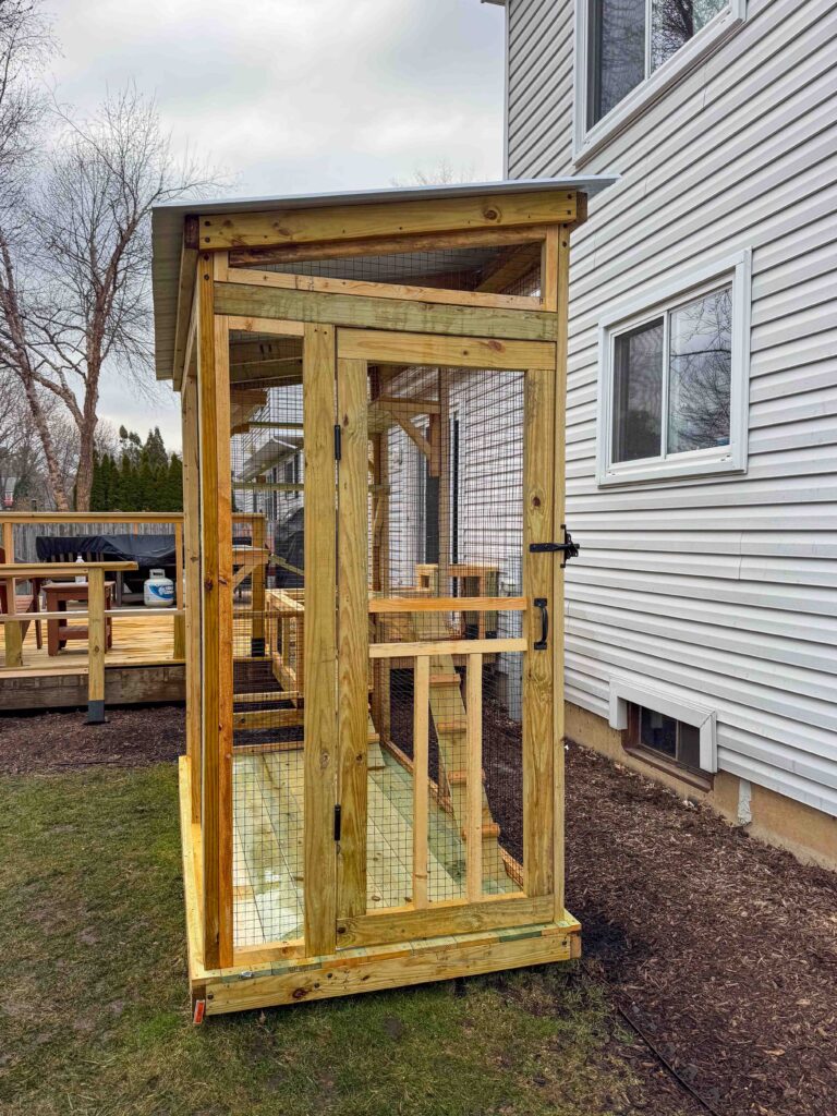 Front-facing view of a custom-built wooden catio in Carol Stream, IL featuring a secure entry door with latch hardware, wire mesh panels, slanted metal roof, and elevated flooring.