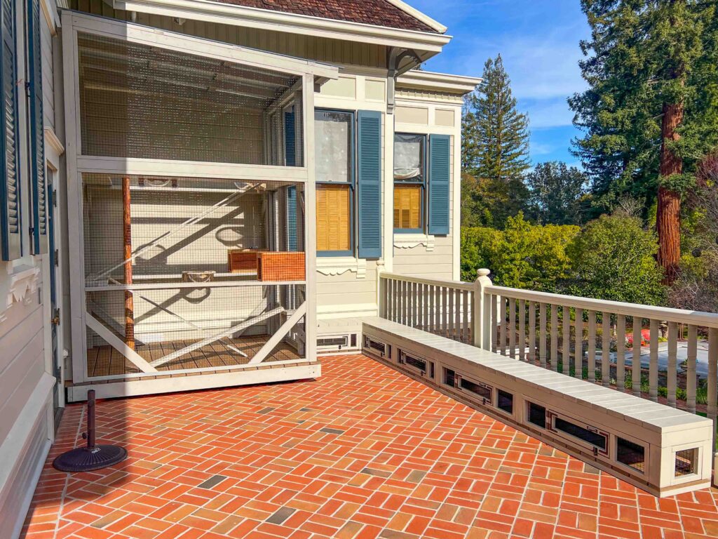Wide-angle view of a custom two-story catio enclosure connected to a Victorian-style home in Atherton, CA, with a matching built-in bench enclosure running along the porch railing and surrounded by mature trees.