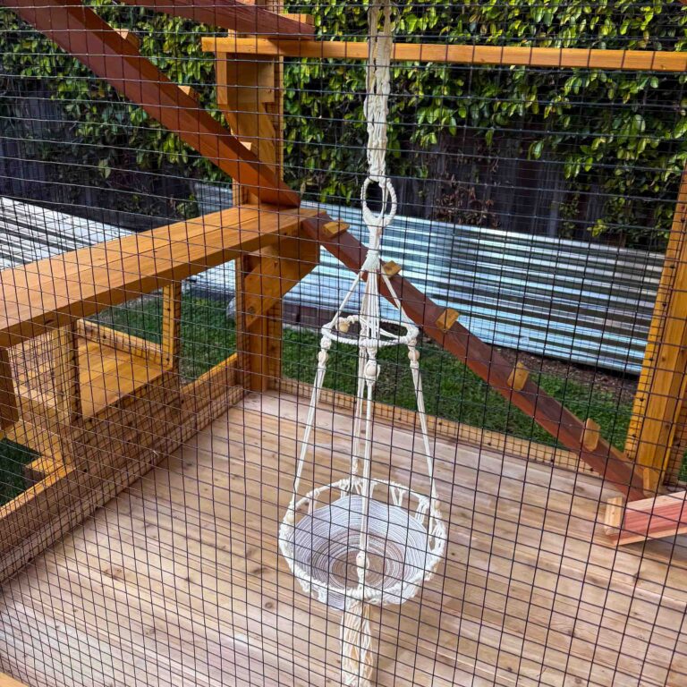 Interior close-up of a wooden catio showing multi-level platforms, an angled climbing ramp, and a suspended rope hammock bed inside a secure mesh enclosure.