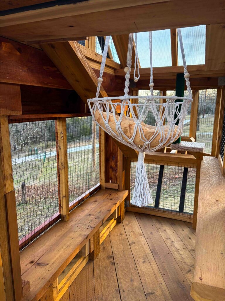 Interior view of custom wooden catio in Aptos, California showcasing natural cedar plank flooring, built-in bench-style shelves, vertical climbing ramp, hanging macramé cat hammock with cushion, wire mesh walls for airflow, and polycarbonate roof panels allowing filtered sunlight.