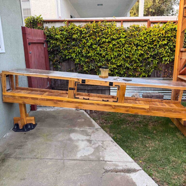 Close-up of an elevated enclosed wooden cat tunnel with wire mesh sides, black trim accents, and sturdy support legs attached to a backyard catio.