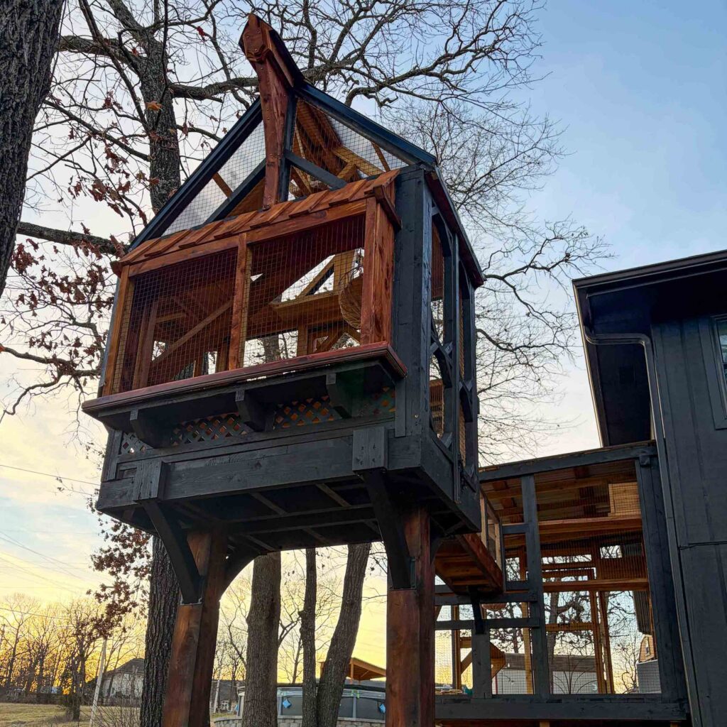 Tall freestanding elevated catio tower with pitched roof, cedar and charcoal wood framing, fully enclosed mesh panels, and sturdy support posts beside a modern dark home exterior at sunset.