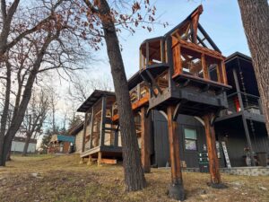 Wide exterior shot of a large elevated multi-level catio attached to a dark modern home, featuring tall wooden support posts, enclosed walkway bridges, and a gabled lookout tower overlooking a sloped yard.