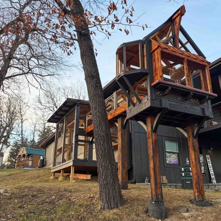 Wide-angle view of multi-level custom catio system attached to home, featuring elevated lookout tower, connecting catwalk bridge, and spacious ground-level enclosure on a sloped wooded property.