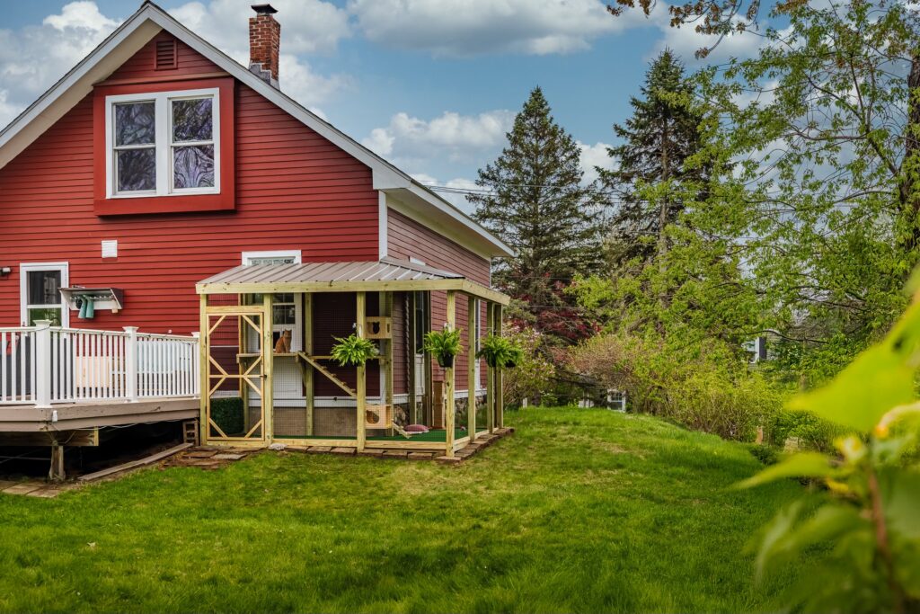 Custom wooden backyard catio attached to a red house in Weston, Massachusetts, featuring a spacious enclosed outdoor cat enclosure with a slanted metal roof, secure wire mesh walls, climbing shelves, ramps, and hanging ferns overlooking a green lawn and trees.